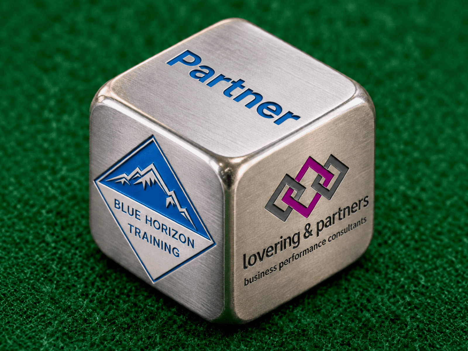 Metal partner cube on green felt, showing the Blue Horizon Training and Lovering & Partners logos